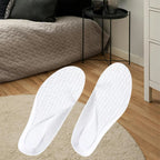 Par de palmilhas ortopédicas X brancas, parea pés chatos, planos, pisada pronada, disposto sobre tapete bege redondo em cenário de quarto com cama ao fundo, destacando a superfície superior texturizada e o design anatômico do calcanhar